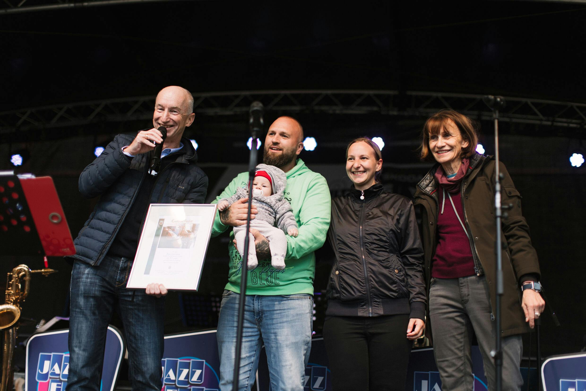 Vier Erwachsene und ein Baby stehen auf einer Bühne. Ein Mann links spricht mit einem Mikrofon und hält eine gerahmte Urkunde in der Hand. Daneben steht ein Mann mit einem Baby im Arm, daneben zwei Frauen, alle lächeln. Im Hintergrund sind Musikinstrumente, Notenständer und ein Banner mit dem Schriftzug Jazz zu sehen. Die Stimmung wirkt festlich und freundlich.