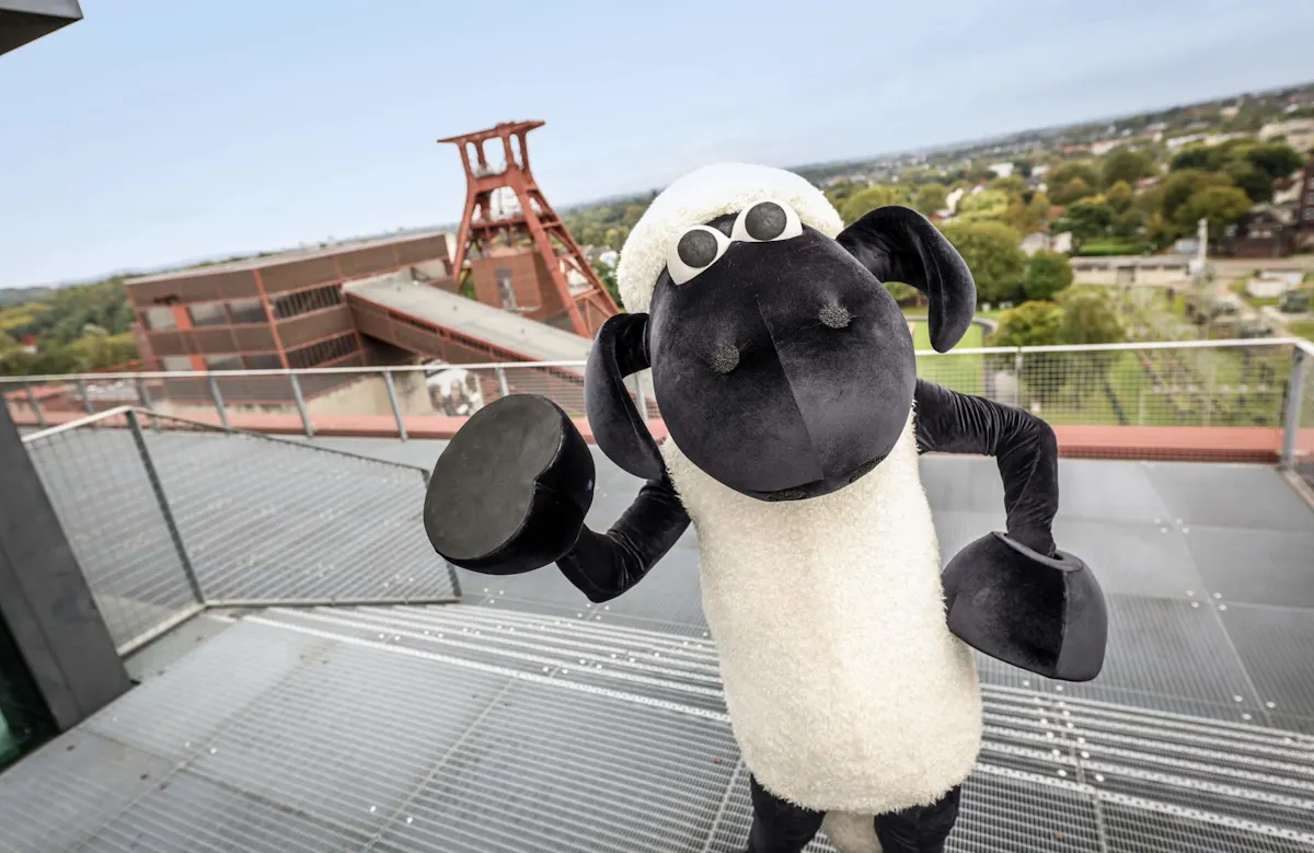 Ein großes Schafmaskottchen mit schwarzem Gesicht und weißen Fell steht auf einer Aussichtsplattform mit Metallgitterboden. Im Hintergrund ist ein Förderturm der Zeche Zollverein in Essen zu sehen, daneben moderne rote Backsteingebäude und eine grüne Parklandschaft. Das Maskottchen hebt einen Arm und scheint fröhlich zu posieren.