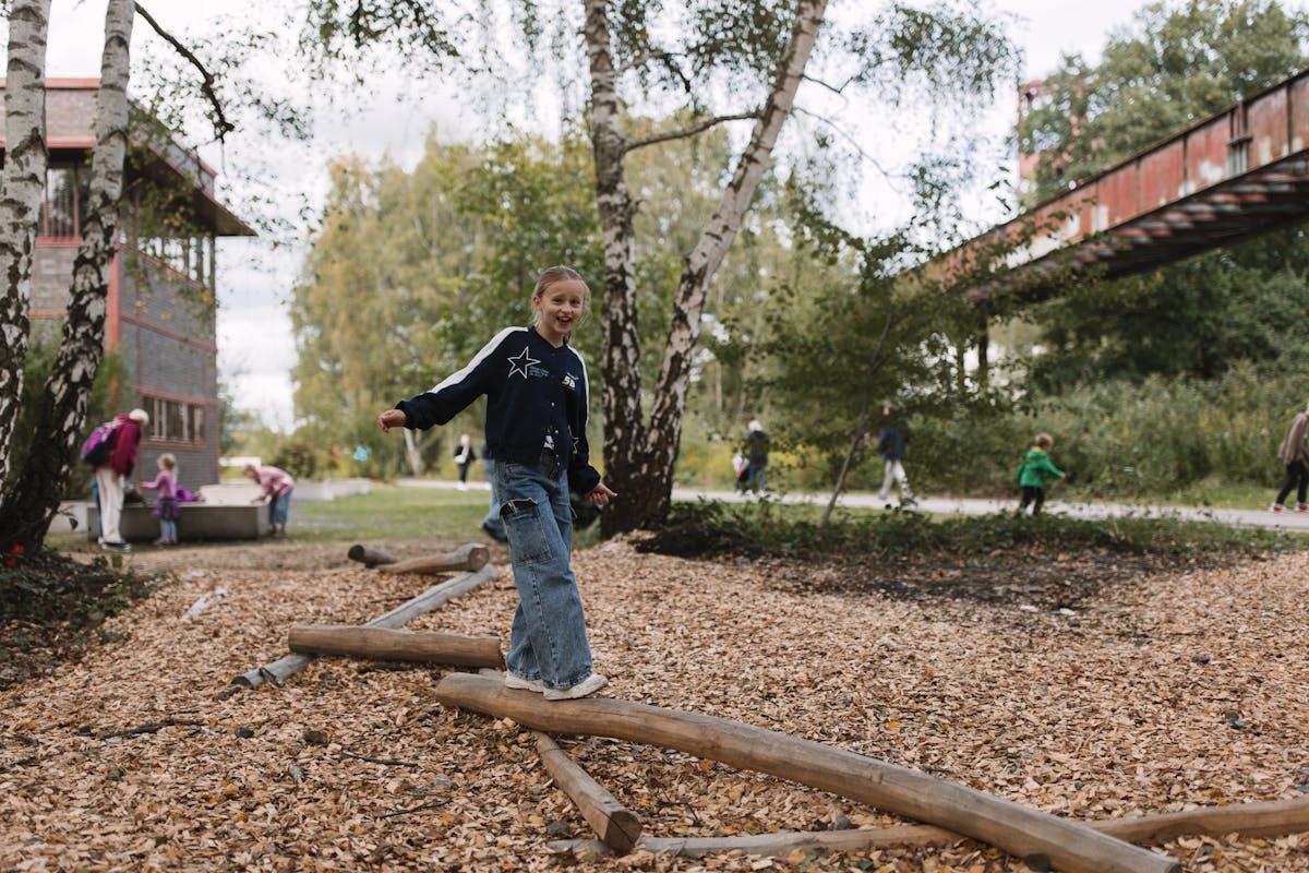 Ein lachendes Mädchen balanciert auf einem Holzstamm in einem mit Rindenmulch ausgelegten Spielbereich zwischen Birkenbäumen. Sie trägt eine dunkelblaue Jacke mit Sternmotiv und weite Jeans. Im Hintergrund sind spielende Kinder, Erwachsene sowie ein rotes Industriegebäude und eine rostige Brücke sichtbar. Die Szene wirkt lebendig und natürlich.