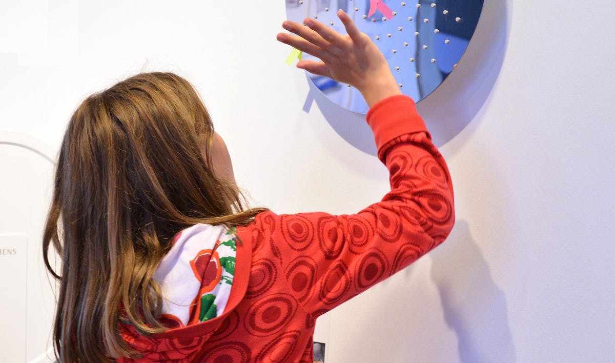 Ein Kind mit langen braunen Haaren und rotem Kapuzenpulli mit Kreismuster hebt die Hand vor einen runden Spiegel mit vielen kleinen Metallstiften. Die Spiegeloberfläche erzeugt verzerrte Lichtreflexionen und farbige Schatten der Hand. Das Kind scheint konzentriert das optische Phänomen zu beobachten.