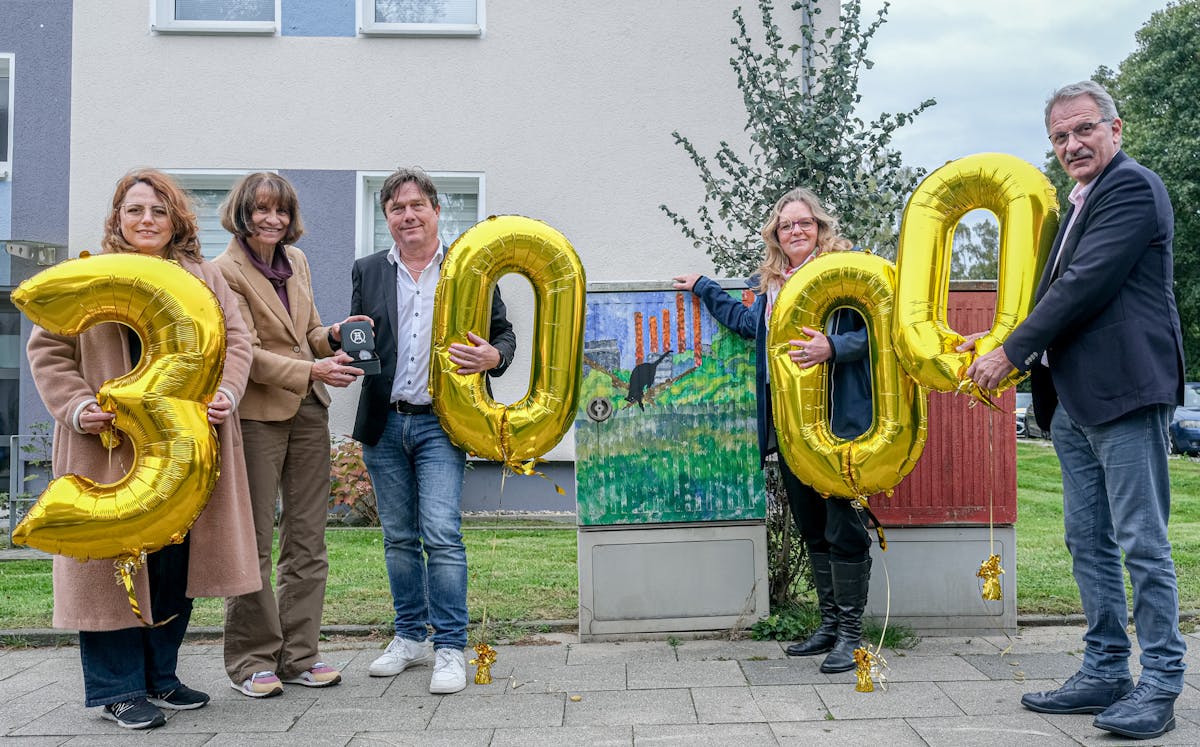 Auf dem Bild sind fünf Personen zu sehen. Sie stehen in einer Reihe und halten goldene Luftballons, die zusammen die Zahl 3000 bilden. In der Mitte befindet sich ein bunt bemalter Stromkasten. Eine der Personen überreicht einer anderen eine kleine Box mit dem Logo der Stiftung Zollverein. Die Stimmung wirkt feierlich.
