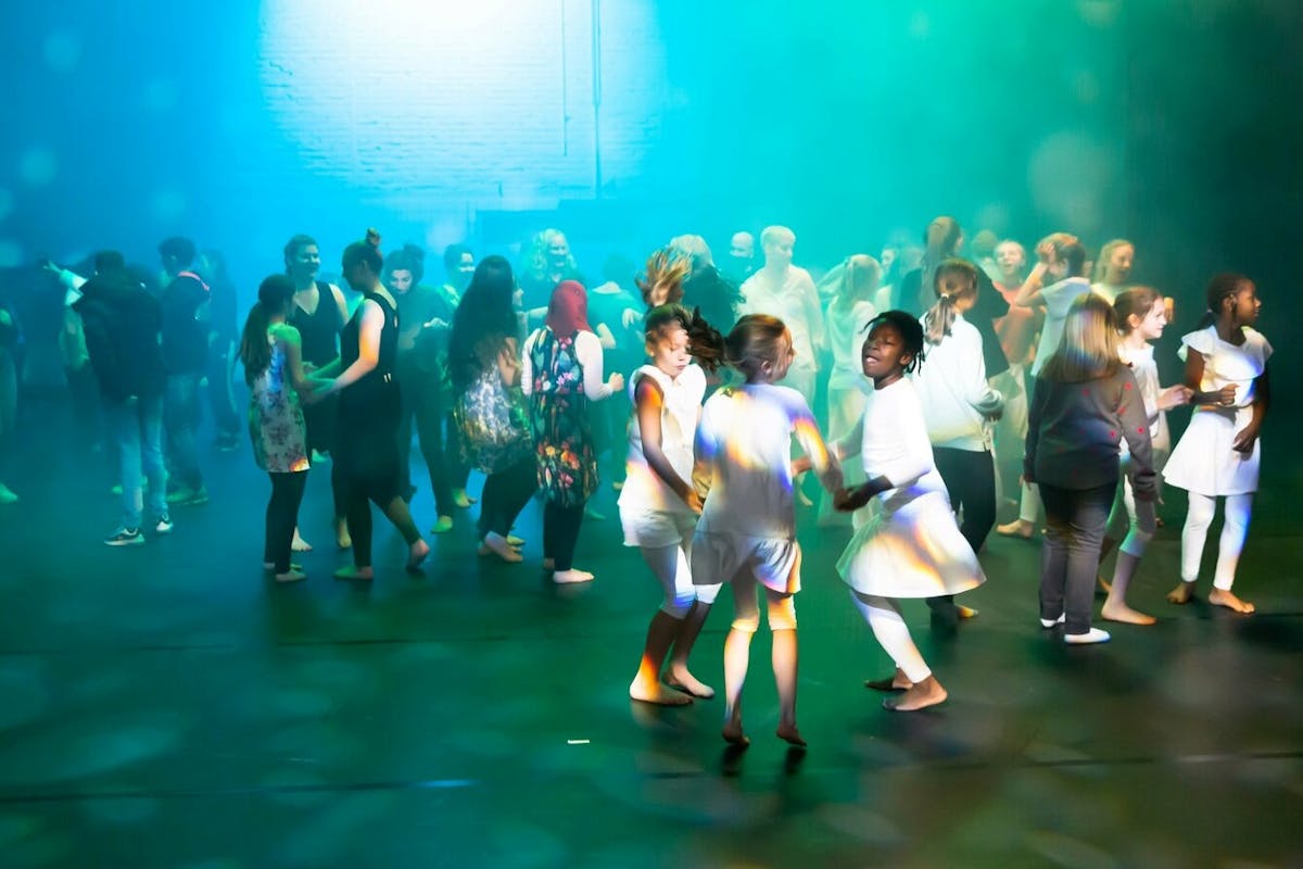 Viele Kinder und Erwachsene tanzen barfuß auf einer dunklen Bühne, die in farbiges Licht und Nebel getaucht ist. Einige Kinder in weißen Kostümen halten sich an den Händen und drehen sich lachend im Kreis. Die Stimmung wirkt lebendig und fröhlich. ChatGPT fragen