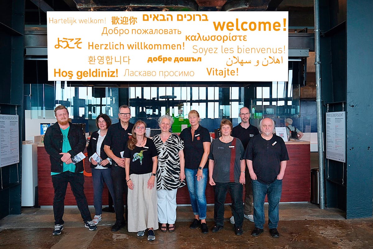 Das Bild zeigt eine Gruppe von zehn Personen, die vor einem Empfangsbereich in einem modernen Gebäude posieren. Sie stehen nebeneinander und lächeln in die Kamera. Einige tragen Namensschilder oder Kleidung mit Logos, was auf ein Team oder Mitarbeitende eines Besucherzentrums hinweist. Über ihnen hängt ein großer Bildschirm mit dem Wort Willkommen in verschiedenen Sprachen, darunter Englisch, Deutsch, Türkisch, Französisch, Arabisch, Hebräisch, Russisch, Chinesisch, Japanisch und viele weitere. Die Atmosphäre wirkt offen, freundlich und einladend. Auf beiden Seiten des Empfangsbereichs hängen Tafeln mit einer Preisübersicht.