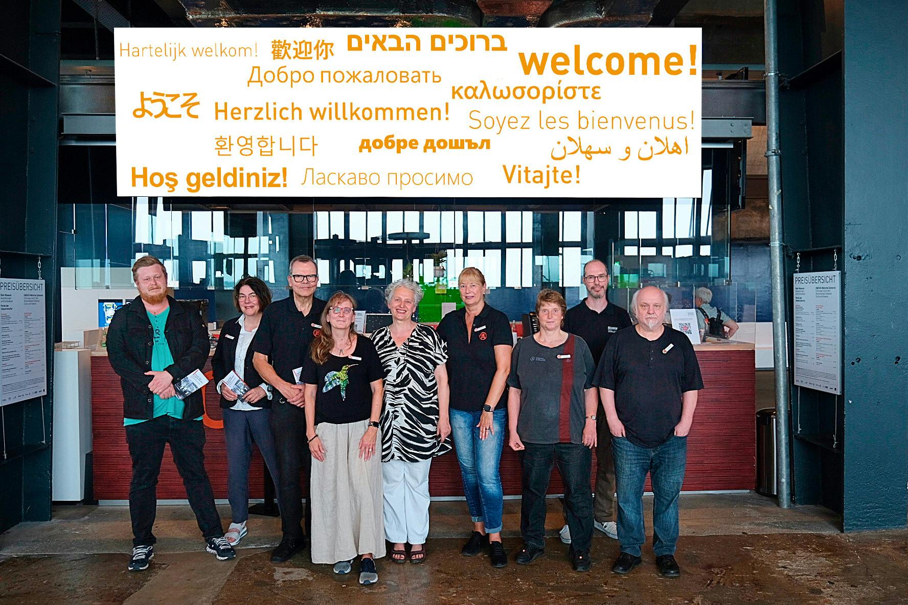 Das Bild zeigt eine Gruppe von zehn Personen, die vor einem Empfangsbereich in einem modernen Gebäude posieren. Sie stehen nebeneinander und lächeln in die Kamera. Einige tragen Namensschilder oder Kleidung mit Logos, was auf ein Team oder Mitarbeitende eines Besucherzentrums hinweist. Über ihnen hängt ein großer Bildschirm mit dem Wort Willkommen in verschiedenen Sprachen, darunter Englisch, Deutsch, Türkisch, Französisch, Arabisch, Hebräisch, Russisch, Chinesisch, Japanisch und viele weitere. Die Atmosphäre wirkt offen, freundlich und einladend. Auf beiden Seiten des Empfangsbereichs hängen Tafeln mit einer Preisübersicht.