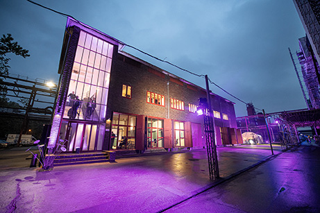 Das Bild zeigt ein Backsteingebäude, im Stil der Neuen Sachlichkeit, auf dem Gelände der Zeche Zollverein in Essen in der Dämmerung oder bei Nacht. Das Gebäude ist von außen durch violette und blaue Lichtquellen effektvoll beleuchtet, was dem Ort eine atmosphärische und kulturelle Ausstrahlung verleiht. Große Fenster an der Frontfassade geben den Blick auf Menschen im Inneren frei. Vor dem Gebäude befindet sich eine nasse, spiegelnde Asphaltfläche, auf der sich das Licht bricht. Metallpfosten mit Lichttechnik und ein überdachter Durchgang auf der rechten Seite deuten auf eine Veranstaltung oder Ausstellung hin. Der Himmel ist bewölkt, was die Lichtstimmung zusätzlich verstärkt.
