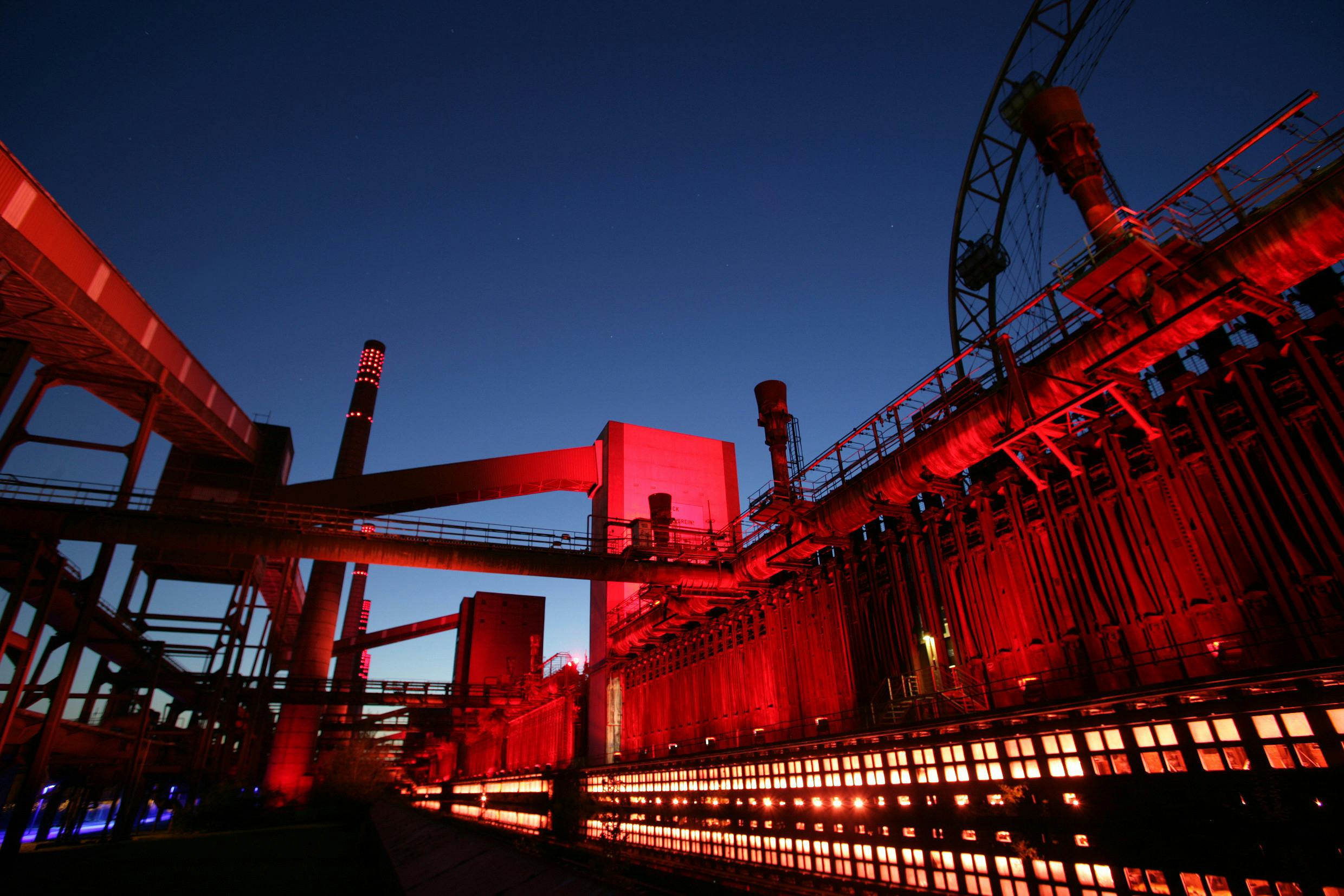 Das Bild zeigt das Industriegelände der Kokerei Zollverein bei Nacht. Die massiven Rohrleitungen, Türme und Industrieanlagen sind in intensivem Rotlicht angestrahlt, was eine dramatische und eindrucksvolle Atmosphäre erzeugt. Am unteren Bildrand leuchten Reihen von Fenstern oder Öffnungen in warmem Orange, was an glühende Ofenkammern erinnert. Der Himmel ist tiefblau und wolkenlos, die Silhouetten der Anlage heben sich klar gegen den Abendhimmel ab. Die Szene wirkt technisch, kraftvoll und kunstvoll inszeniert.