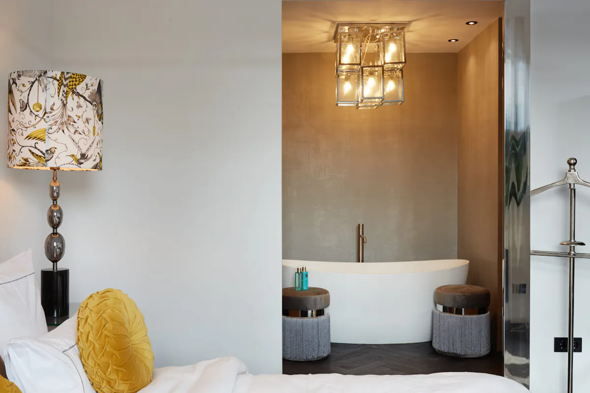 Das Bild zeigt ein stilvoll eingerichtetes Hotelzimmer mit direktem Blick auf eine freistehende Badewanne. Im Vordergrund links befindet sich ein Bett mit weißer Bettwäsche und einem runden, senfgelben Zierkissen. Daneben steht eine Tischlampe mit einem auffällig gemusterten Lampenschirm mit Vogelmotiven in Gelb, Schwarz und Grau. Im Hintergrund ist die weiße, ovale Badewanne in einer Nische mit goldfarbenen Wänden platziert, flankiert von zwei grauen Hockern mit Fransen. Eine moderne Deckenleuchte mit mehreren Glaskuben erhellt den Raum. Rechts im Bild ist ein glänzender Kleiderständer sichtbar. Das Gesamtbild vermittelt einen Eindruck von modernem Luxus mit einem Hauch Retro-Charme.