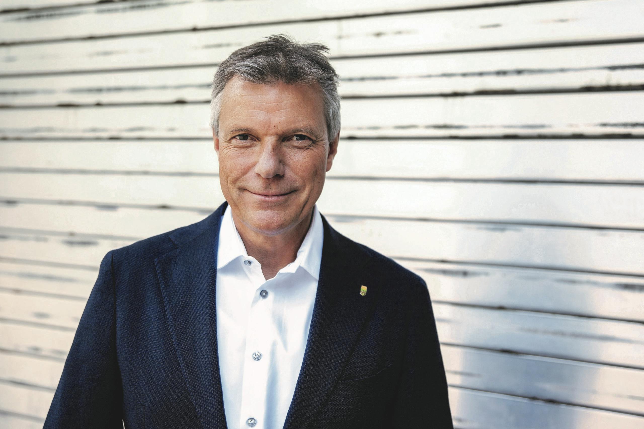 Ein Mann mittleren Alters mit grauen Haaren steht lächelnd vor einer weißen, horizontalen Holzverkleidung. Er trägt ein dunkelblaues Sakko über einem weißen Hemd und schaut direkt in die Kamera. Die Atmosphäre ist freundlich und professionell.