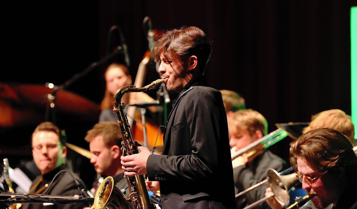 Ein junger Musiker steht im Vordergrund und spielt konzentriert Saxofon, umgeben von weiteren Musikerinnen und Musikern in einem Orchester. Alle tragen dunkle Kleidung und sind auf ihre Instrumente fokussiert. Im Hintergrund sind Trompeten, Posaunen und ein Kontrabass zu erkennen, ebenso wie Mikrofone, die auf die Gruppe gerichtet sind. Die Szene spielt sich auf einer Bühne mit dunklem Hintergrund ab, die Beleuchtung hebt die Gesichter und Instrumente farbenfroh hervor.
