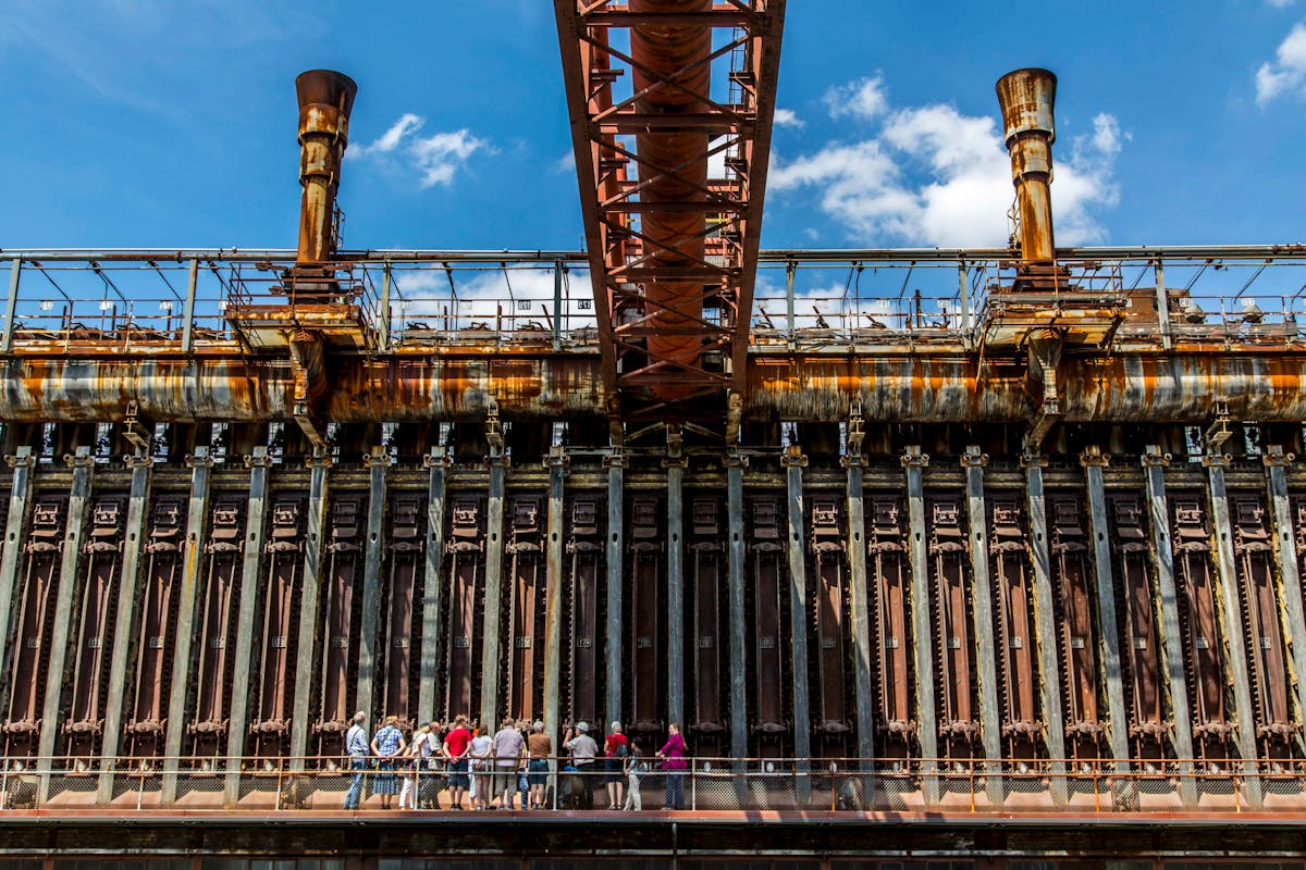 Eine Gruppe von Menschen steht auf einer Besucherplattform vor einer monumentalen, rostigen Industrieanlage mit zahlreichen vertikalen Metalltüren und Röhren. Oberhalb verlaufen rostige Rohre und Gitterstrukturen, in der Mitte ragt eine große, rostige Rohrverbindung horizontal hervor. Der blaue Himmel mit einigen Wolken bildet einen starken Kontrast zur verwitterten und dunklen Fassade der Anlage. Die Szene vermittelt den Eindruck einer Führung durch ein stillgelegtes Industrieareal.