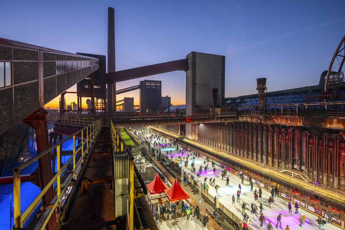 Abendaufnahme eines ehemaligen Industrieareals mit beleuchteter Eislaufbahn im Zentrum, auf der zahlreiche Menschen Schlittschuh laufen. Im Hintergrund sind alte Industriegebäude, Förderanlagen und ein hoher Schornstein zu sehen, die in warmes Licht getaucht sind. Über dem Horizont zeigt sich ein farbenfroher Sonnenuntergang, während die Szene durch bunte Beleuchtung lebendig wirkt.