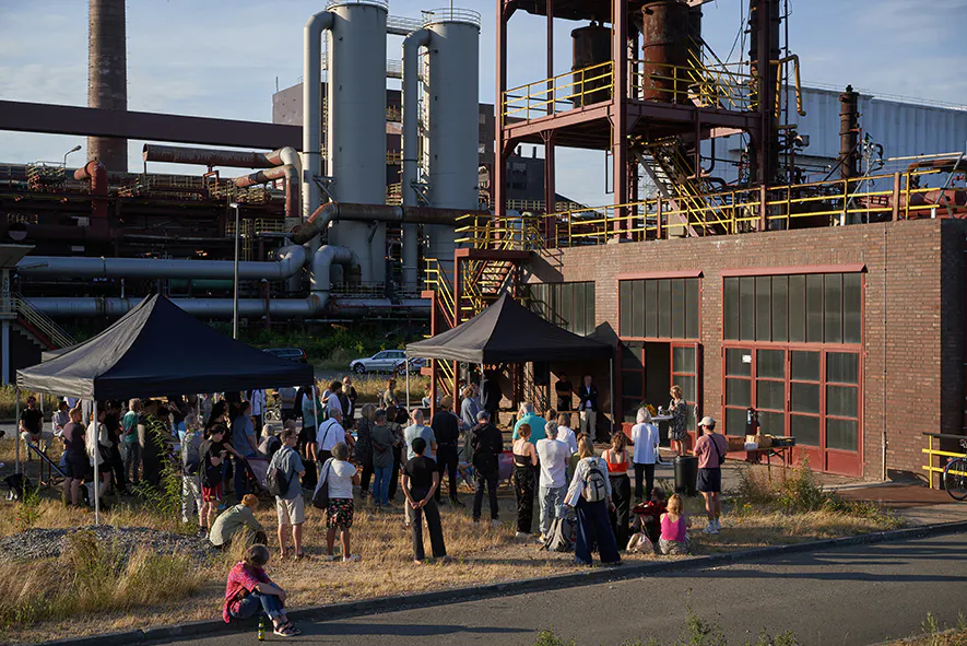 Das Bild zeigt eine Gruppe von Menschen, die sich bei einer Veranstaltung im Freien vor einem alten Industriegebäude mit großen Fenstern versammelt haben. Zwei schwarze Pavillons bieten Schatten für Redner oder Ausstellungsobjekte. Im Hintergrund ragen industrielle Anlagen mit Rohren, Tanks und Stahlkonstruktionen in die Höhe. Einige Personen stehen, andere sitzen im Gras oder auf dem Asphalt. Die Szene spielt bei sonnigem Wetter und vermittelt eine Atmosphäre von Kultur oder Bildung inmitten industrieller Kulisse.
