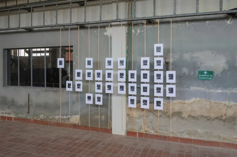 Das Bild zeigt eine Wandinstallation in einem alten Industriegebäude mit abblätterndem Putz und freiliegenden Leitungen. An dünnen Schnüren sind zahlreiche weiße Papierbögen aufgehängt, die jeweils ein kleines, zentriertes schwarz-weißes Foto zeigen. Die Anordnung der Bögen bildet eine gleichmäßige rechteckige Fläche. Links befindet sich ein Fenster mit Blick in einen weiteren Raum, rechts ein grünes Schild mit der Aufschrift Hier befindet sich eine Krankentrage. Der Boden besteht aus roten Fliesen, die Szene wirkt künstlerisch, provisorisch und dokumentarisch zugleich.