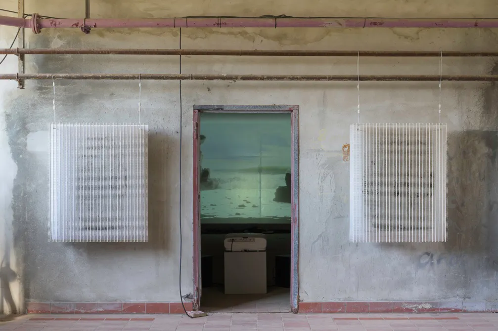 Das Bild zeigt einen Raum mit rohen, grauen Wänden und offenen Rohrleitungen unter der Decke. Links und rechts neben einer Türöffnung hängen zwei große, filigrane Objekte aus weißen, vertikal ausgerichteten Stäben. Durch die offene Tür ist ein abgedunkelter Raum zu sehen, in dem an der Rückwand ein Video mit einer Landschaftsaufnahme, vermutlich einer Meeres- oder Küstenszene, projiziert wird. Im Vordergrund steht ein Gerät, das wie ein Projektor oder Drucker aussieht. Die Szene wirkt minimalistisch und technisch, mit einem Kontrast zwischen Struktur und Naturbild.