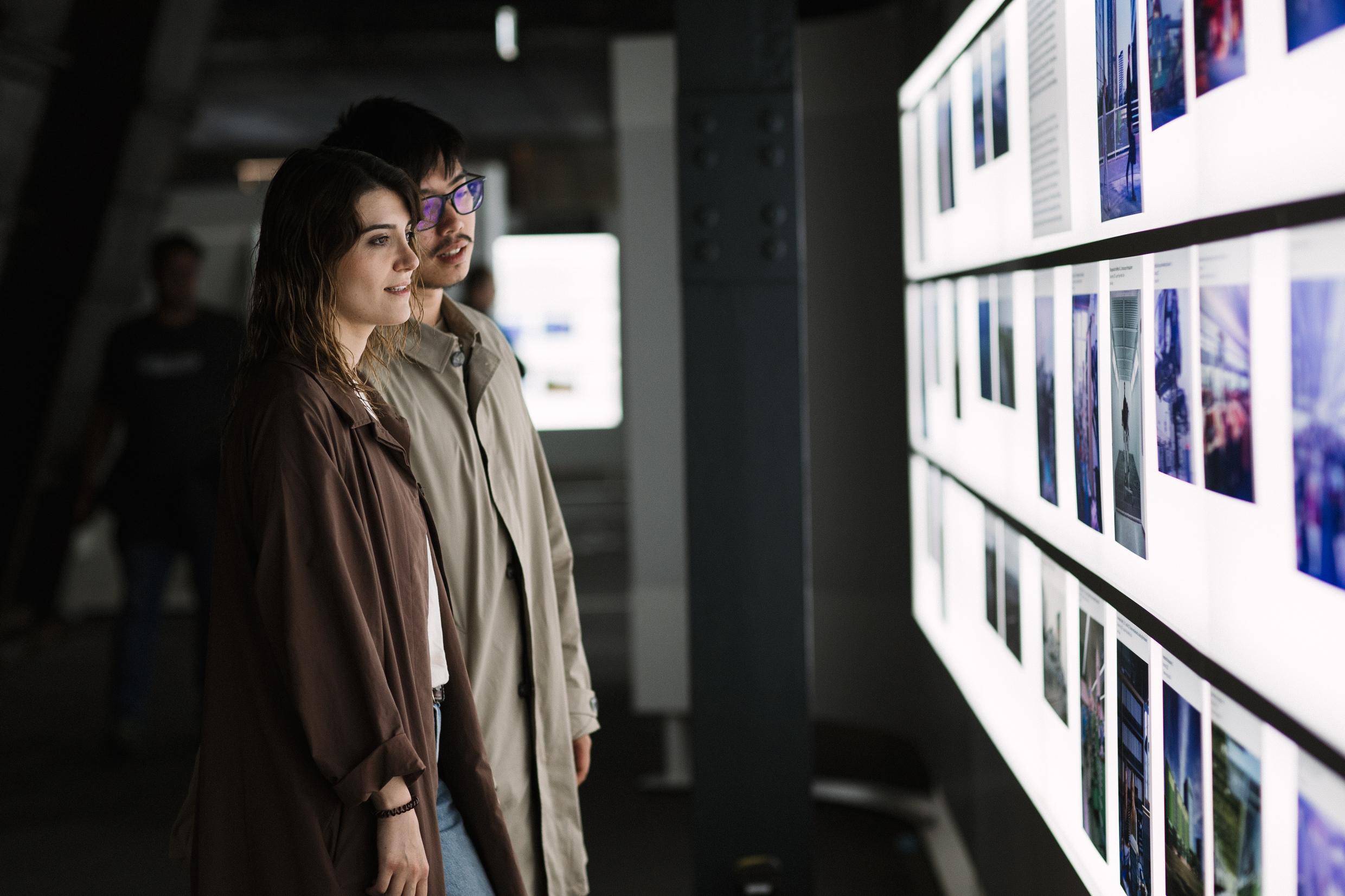 Ein junges Paar steht nebeneinander in einem abgedunkelten Ausstellungsraum und betrachtet interessiert eine beleuchtete Wand mit vielen kleinen Fotos und Texten. Die Frau trägt einen braunen Mantel, der Mann einen beigefarbenen Trenchcoat und eine Brille mit lilafarbenen Gläsern. Im Hintergrund sind weitere Besucher schemenhaft zu erkennen.