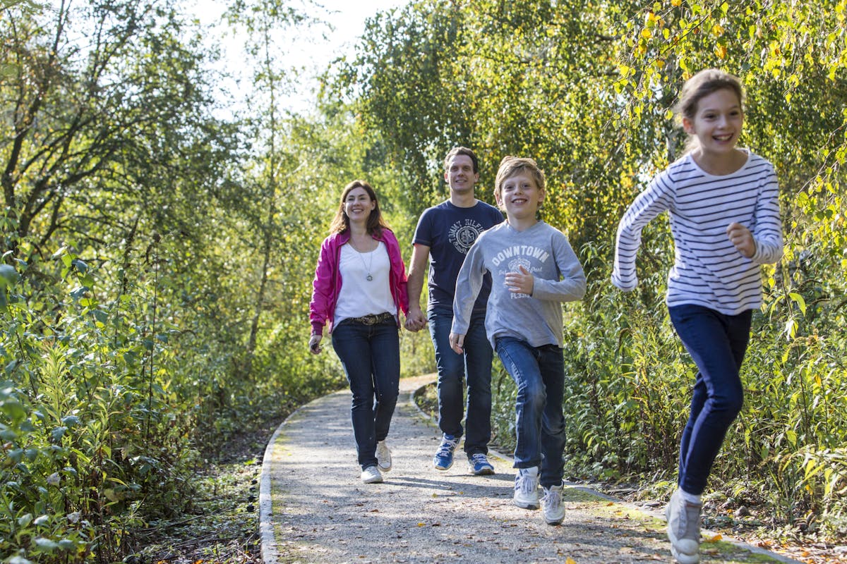Eine fröhliche Familie ist auf einem schmalen Waldweg unterwegs. Zwei Kinder rennen lachend im Vordergrund – ein Mädchen im gestreiften Shirt und ein Junge im grauen Pullover. Dahinter gehen die Eltern Hand in Hand, die Mutter trägt eine pinke Jacke und der Vater ein dunkelblaues T-Shirt. Die Umgebung ist grün und sonnig, mit dichtem Laub an beiden Seiten des Weges. Die Szene vermittelt Freude, Bewegung und gemeinsames Erleben in der Natur.