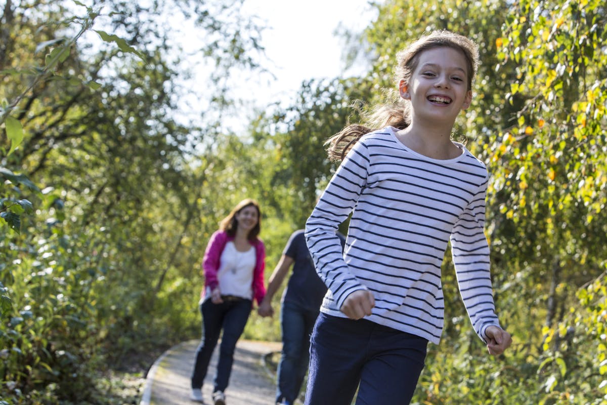 Ein junges Mädchen mit einem weiß-blau gestreiften Langarmshirt läuft fröhlich auf einem schmalen Weg durch einen grünen, sonnendurchfluteten Wald. Im Hintergrund gehen zwei Erwachsene, eine Frau in einer pinken Jacke und ein Mann, Hand in Hand. Die Szene wirkt lebendig und zeigt einen unbeschwerten Moment in der Natur.