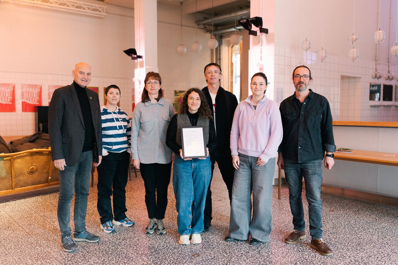 Das Bild zeigt eine Gruppe von sieben Personen, die in einem hellen, modern eingerichteten Raum nebeneinander stehen und in die Kamera lächeln. In der Mitte hält eine Frau ein gerahmtes Dokument in den Händen. Die Personen sind leger bis leicht formell gekleidet. Der Raum hat weiße Wände mit Fliesen, Hängelampen und ein paar rote Poster an der Wand. Links im Hintergrund ist eine goldfarbene Couch zu sehen, rechts eine Theke mit Holzplatte. Die Atmosphäre wirkt freundlich und entspannt, möglicherweise handelt es sich um eine Preisverleihung oder die Präsentation eines Projektergebnisses.