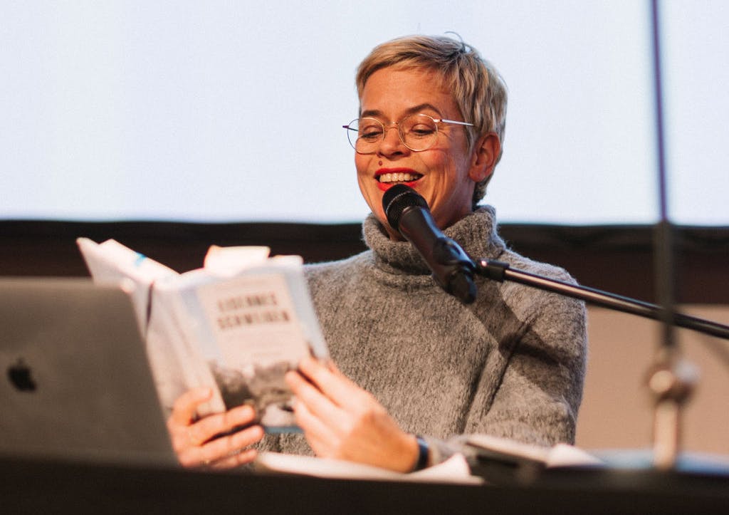 Eine lächelnde Frau mit kurzem blondem Haar und Brille sitzt an einem Tisch und liest aus einem Buch mit dem Titel Eisernes Schweigen. Sie trägt einen grauen Rollkragenpullover und spricht gleichzeitig in ein Mikrofon. Vor ihr stehen ein aufgeklappter Laptop und weitere Unterlagen. Die Szene wirkt wie eine öffentliche Lesung oder ein Vortrag in einem hell beleuchteten Raum.