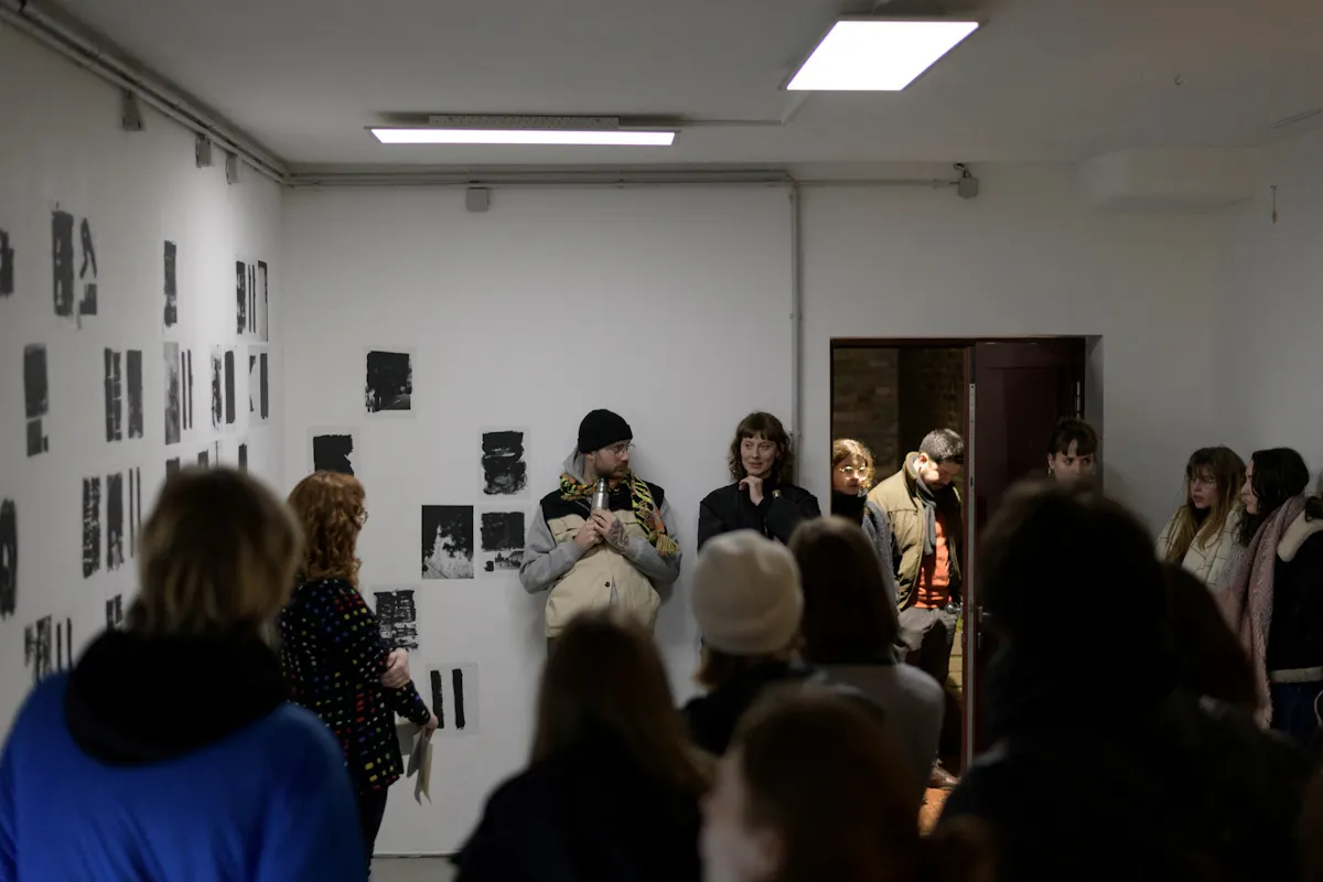 Ein Gruppenfoto zeigt eine Szene in einem kleinen, weißen Raum mit Kunstwerken an der linken Wand, die aus abstrakten Schwarz-Weiß-Drucken bestehen. Eine Gruppe von Menschen steht und sitzt dicht gedrängt im Raum, einige hören aufmerksam zu, andere unterhalten sich. Im Fokus stehen zwei Personen vor der Kunstwand – ein Mann mit Mütze, Brille und Thermoskanne sowie eine Frau mit dunklen Haaren und schwarzer Kleidung. Sie wirken wie Gastgeber oder Vortragende. Im Hintergrund steht eine weitere Gruppe von Menschen in einem offenen Türrahmen, teilweise im Schatten. Die Beleuchtung ist überwiegend künstlich und erzeugt eine konzentrierte, intime Atmosphäre, wie bei einer Ausstellungseröffnung oder Künstlerpräsentation.