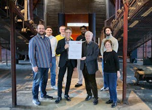 Eine Gruppe von neun Personen steht im Innenraum eines historischen Industriegebäudes mit metallenen Treppen und Stahlträgern. Zwei Männer in der Mitte halten gemeinsam eine offizielle Urkunde in die Kamera, sie lächeln stolz. Im Hintergrund ist ein Schild mit der Aufschrift Ruhr Museum zu sehen. Die Personen wirken fröhlich und feiern offenbar eine Auszeichnung oder ein besonderes Ereignis.