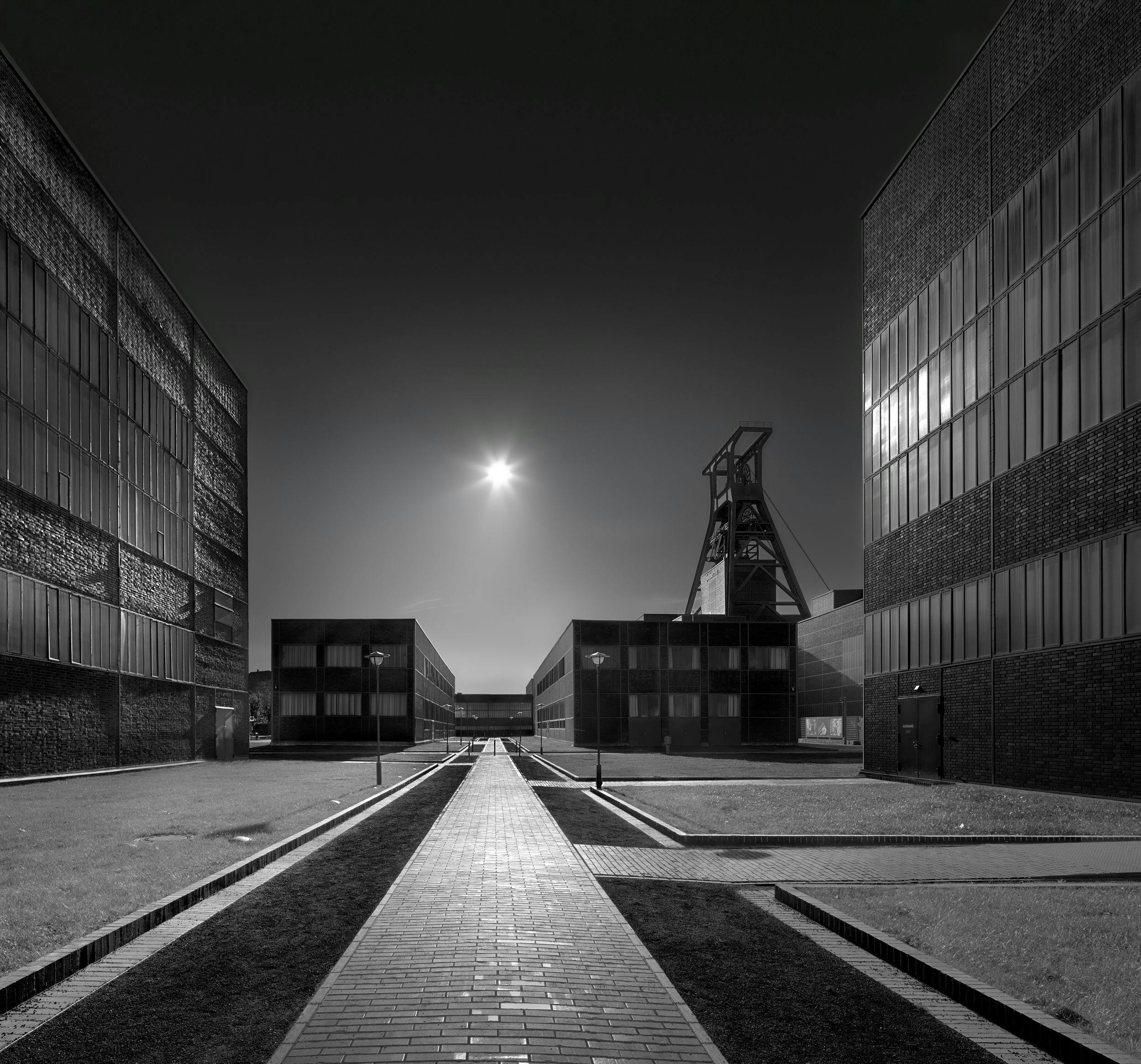 Schwarz-weiß-Fotografie eines symmetrischen Industrieareals bei Nacht oder in der Dämmerung. Im Vordergrund führt ein gepflasterter Weg mittig durch gepflegte Rasenflächen, gesäumt von Straßenlaternen. Rechts und links stehen moderne Backsteingebäude mit großen Fensterflächen. Im Hintergrund ragt zentral ein historischer Förderturm empor, beleuchtet von einem hellen Lichtpunkt am Himmel, vermutlich die Sonne. Die Szene wirkt ruhig, klar und architektonisch streng.