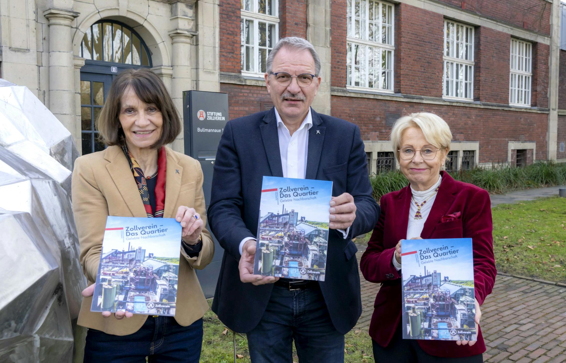 Drei Personen stehen vor einem historischen Backsteingebäude mit großen Fenstern und halten jeweils ein Exemplar einer Broschüre mit dem Titel Zollverein – Das Quartier Gelebte Nachbarschaft in die Kamera. Die Frau links trägt ein beigefarbenes Jackett, der Mann in der Mitte einen dunkelblauen Anzug und die Frau rechts ein rotes Samtjackett. Im Hintergrund ist ein Schild mit der Aufschrift Stiftung Zollverein und der Straßenname Bullmannaue zu sehen. Alle drei Personen lächeln und präsentieren stolz die Publikation.