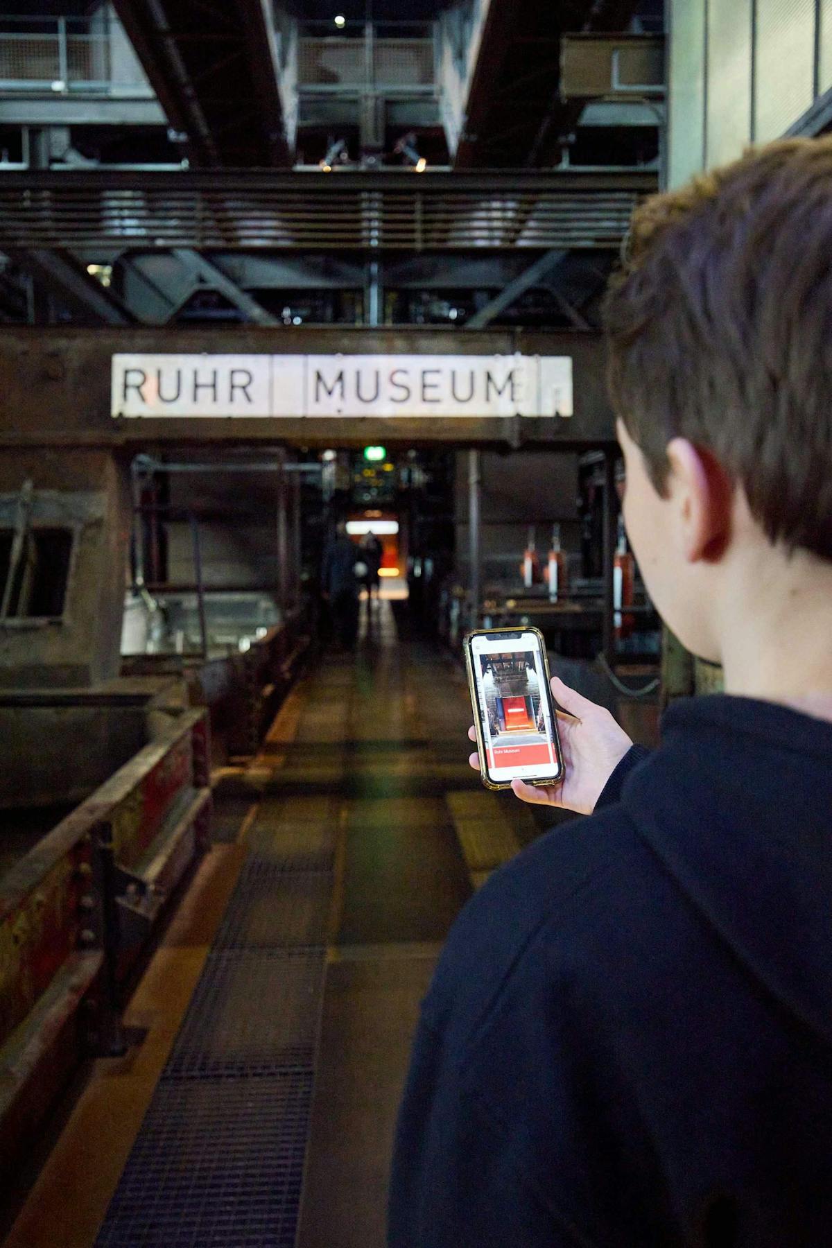 Ein junger Mann steht in einem industriellen Gebäude vor einem beleuchteten Schild mit der Aufschrift Ruhr Museum. Er hält ein Smartphone in der Hand, auf dessen Bildschirm dieselbe Szene mit dem Museumseingang angezeigt wird. Die Umgebung ist dunkel und besteht aus Metallstrukturen und Laufstegen, was auf eine frühere industrielle Nutzung des Gebäudes hindeutet.