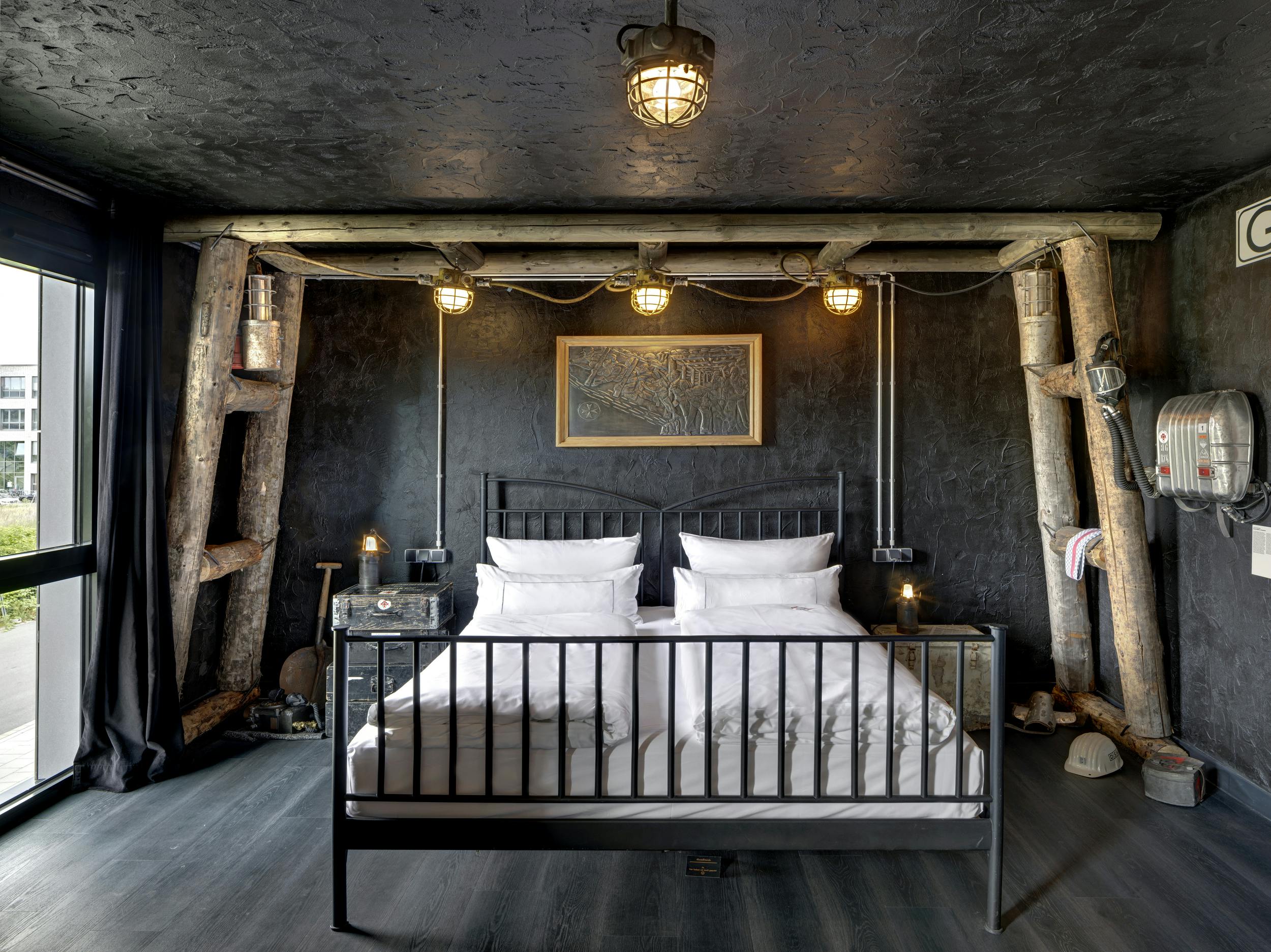 Ein stilvoll eingerichtetes Schlafzimmer mit industriellem und rustikalem Flair. In der Mitte steht ein schwarzes Metallbett mit weißer Bettwäsche. Die Wände und die Decke sind in dunklem Grau verputzt, der Boden ist aus dunklem Holz. Über dem Bett hängen vier rustikale Lampen, befestigt an einer Konstruktion aus groben Holzstämmen. Links und rechts vom Bett befinden sich Nachttische mit kleinen Lampen. Die Wand ist mit einer Zeichnung dekoriert und links ist ein Fenster mit schwarzem Vorhang. Bergbauhelme, Werkzeuge und ein Atemschutzgerät am Boden und an der Wand verleihen dem Raum eine bergwerkartige Atmosphäre.