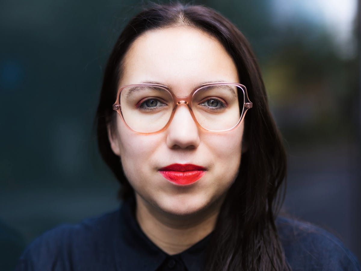 Porträtaufnahme einer Frau mit heller Haut, dunklen, langen Haaren und auffälligem roten Lippenstift. Sie trägt eine große, transparente Brille mit rosafarbenem Rahmen und ein dunkles Hemd. Die Frau blickt direkt in die Kamera mit einem neutralen Gesichtsausdruck. Der Hintergrund ist unscharf und dunkel, wodurch der Fokus ganz auf dem Gesicht liegt.