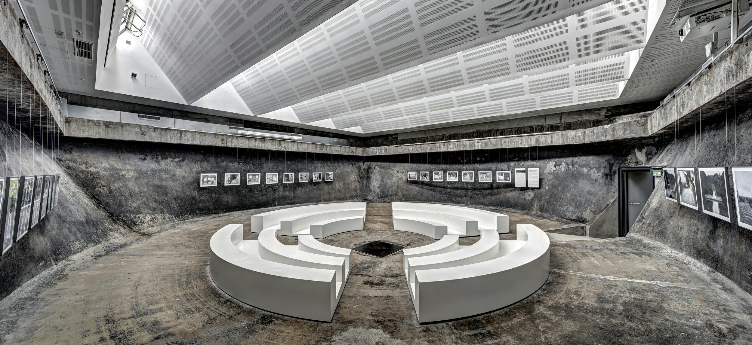 Futuristisch gestalteter Raum mit kreisförmigen weißen Sitzbänken im Zentrum, die wie ein Amphitheater angeordnet sind. Der Boden und die Wände bestehen aus rauem Beton, was dem Raum ein industrielles Aussehen verleiht. Rund um den Raum hängen gerahmte Schwarz-Weiß-Fotografien an Drahtseilen von der Decke. Der helle, geometrisch strukturierte Deckenbereich steht im starken Kontrast zur dunklen Umgebung.