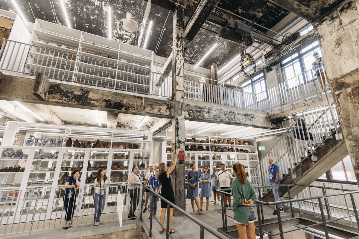 Das Bild zeigt den Innenraum eines Museums oder Depots mit industriellem Charakter. Die Architektur ist geprägt von rohem Beton, sichtbaren Stahlträgern und einer offenen Galerie mit Metallgeländern. Auf mehreren Ebenen sind weiße Regale mit zahlreichen Keramikobjekten bestückt, darunter Vasen, Töpfe und Schalen in verschiedenen Formen und Farben. Besucherinnen und Besucher bewegen sich durch den Raum, einige stehen in Gruppen zusammen, andere hören einer Frau zu, die anscheinend eine Führung leitet. Die Atmosphäre ist hell, modern und lebendig, mit einer gelungenen Verbindung aus historischer Bausubstanz und zeitgemäßer Präsentation.