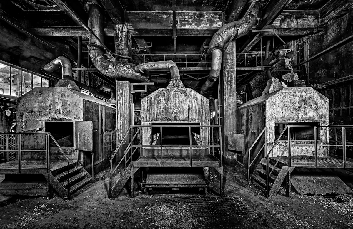 Schwarz-weiß-Fotografie einer verlassenen Industrieanlage mit drei großen, stark verrosteten Ofen- oder Brennkammern, die nebeneinander stehen. Jeder Ofen hat eine kleine Metalltreppe und eine Plattform mit Geländer davor. Die Türen der Brennkammern sind teilweise geöffnet. Über den Öfen verlaufen dicke, verwitterte Rohrleitungen und Metallträger, die die Decke stützen. Die Umgebung ist stark verschmutzt, die Oberflächen wirken abgenutzt und von der Zeit gezeichnet. Die Szenerie vermittelt eine düstere, verlassene Atmosphäre eines früheren industriellen Nutzraums.