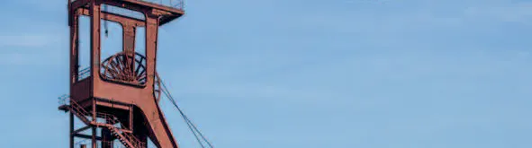 Das Bild zeigt das Titelmotiv eines Flyers oder Plakats für das UNESCO-Welterbe Zollverein. Im oberen Bereich steht in schwarzer Schrift auf weißem Hintergrund: Einsteigen bitte! E-Busfahrten über Zeche und Kokerei. Rechts daneben befindet sich ein roter Kreis mit der Jahreszahl 2025. Links am Rand verläuft vertikal der Schriftzug UNESCO-Welterbe Zollverein. Im unteren Bildteil ist das bekannte rote Fördergerüst der Zeche Zollverein zu sehen, umgeben von Bäumen und grünem Bewuchs. Davor fahren zwei kleine weiße E-Busse mit offenen Seiten, in denen Besucherinnen und Besucher sitzen. Unten rechts befindet sich das Logo der Stiftung Zollverein. Die Szene vermittelt eine Einladung zu einer umweltfreundlichen Entdeckungstour über das ehemalige Industrieareal.