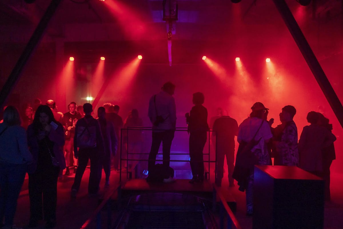 Zahlreiche Besucherinnen und Besucher stehen in rotem Licht und Nebel in der Mischanlage auf dem Zollverein-Gelände. Die Atmosphäre ist dicht und clubähnlich, einzelne Personen unterhalten sich oder betrachten den Raum.