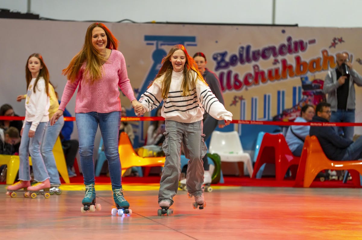 In einer bunten Rollschuhhalle fahren mehrere Menschen fröhlich auf Rollschuhen. Im Vordergrund halten sich eine Frau im pinken Pullover und ein Mädchen im gestreiften Sweatshirt an den Händen, beide lachen und bewegen sich dynamisch. Im Hintergrund sind weitere Kinder und Erwachsene auf Rollschuhen sowie farbenfrohe Kunststoffstühle und Zuschauer zu sehen. An der Wand steht in großen Buchstaben Zollverein-Rollschuhbahn. Die Szene wirkt lebendig und vermittelt Spaß an Bewegung und Gemeinschaft.