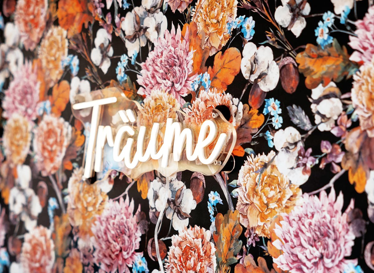 Detailaufnahme einer bunten Blumenmuster-Tapete mit großflächigen Blüten in Rosa, Orange, Weiß und Blau sowie herbstlich gefärbten Blättern auf dunklem Hintergrund. Im Zentrum leuchtet das Wort Träume in heller Schreibschrift als Neon-Schriftzug. Ein transparenter Stromkabel verläuft nach unten. Die Szene wirkt verträumt, kreativ und dekorativ.