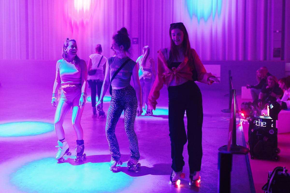 Drei junge Frauen fahren lachend auf Rollschuhen in einer Halle mit bunter Lichtstimmung. Die Szenerie ist in kräftigem Rosa-, Blau- und Violettlicht getaucht, was eine lebendige und festliche Atmosphäre erzeugt. Alle drei tragen auffällige, modische Kleidung, darunter ein rosa Zweiteiler, ein Leopardenmuster und eine rote Bluse. Sie bewegen sich gemeinsam über die Fläche und genießen den Moment. Im Hintergrund sitzen einige Personen auf weißen Sitzwürfeln und beobachten das Geschehen. Die Umgebung erinnert an eine Rollschuhdiskothek mit Partystimmung.