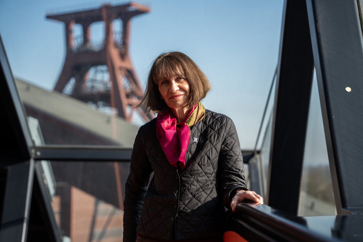 Eine Frau mittleren Alters mit braunen, schulterlangen Haaren steht lächelnd auf einer verglasten Brücke oder Galerie. Sie trägt eine gesteppte schwarze Jacke und ein farbenfrohes Halstuch in Rot und Grün. Im Hintergrund ist unscharf das markante Fördergerüst der Zeche Zollverein bei blauem Himmel zu erkennen. Die Szene wirkt freundlich, selbstbewusst und verbindet Mensch mit Industriekultur.