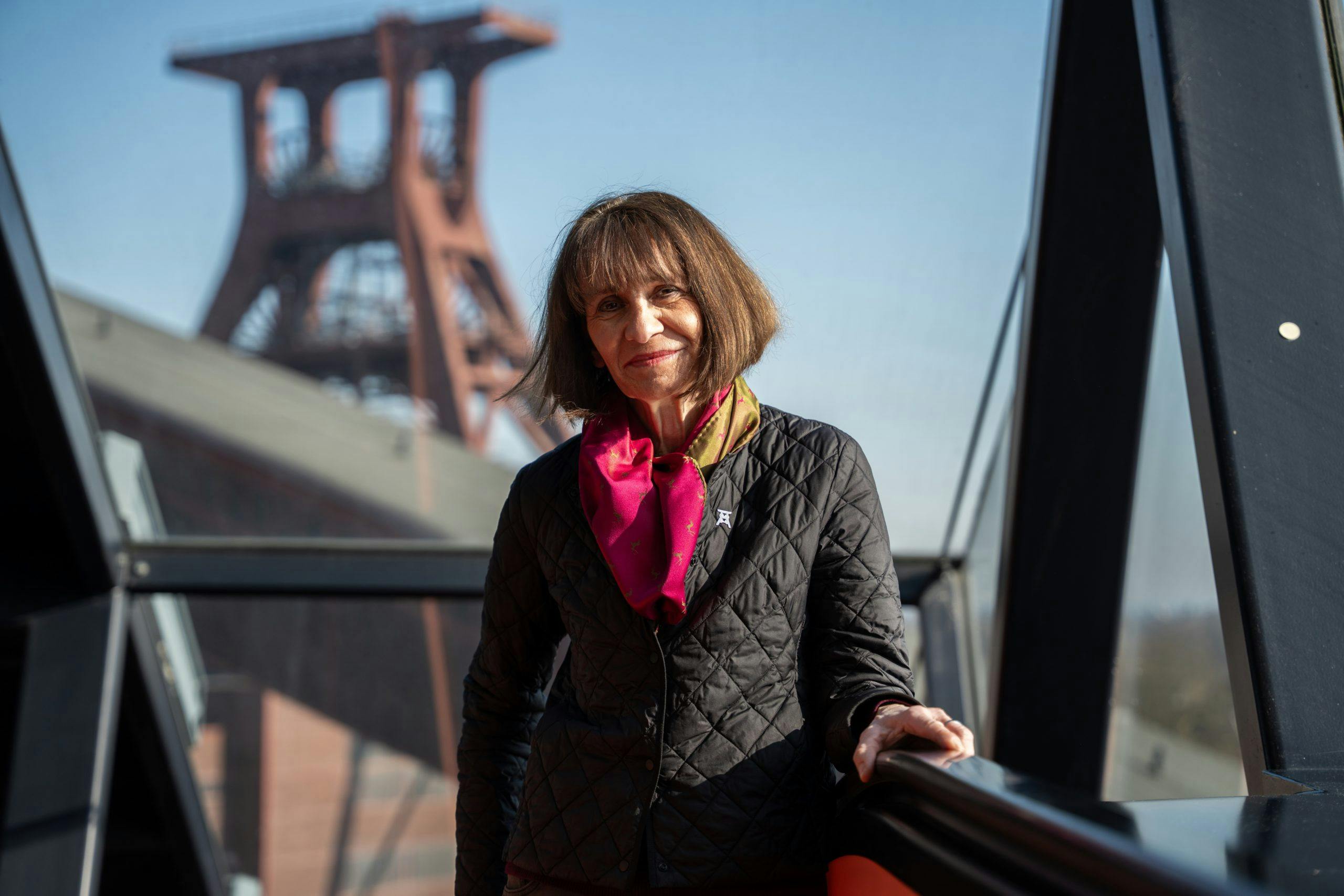 Eine Frau mittleren Alters mit braunen, schulterlangen Haaren steht lächelnd auf einer verglasten Brücke oder Galerie. Sie trägt eine gesteppte schwarze Jacke und ein farbenfrohes Halstuch in Rot und Grün. Im Hintergrund ist unscharf das markante Fördergerüst der Zeche Zollverein bei blauem Himmel zu erkennen. Die Szene wirkt freundlich, selbstbewusst und verbindet Mensch mit Industriekultur.