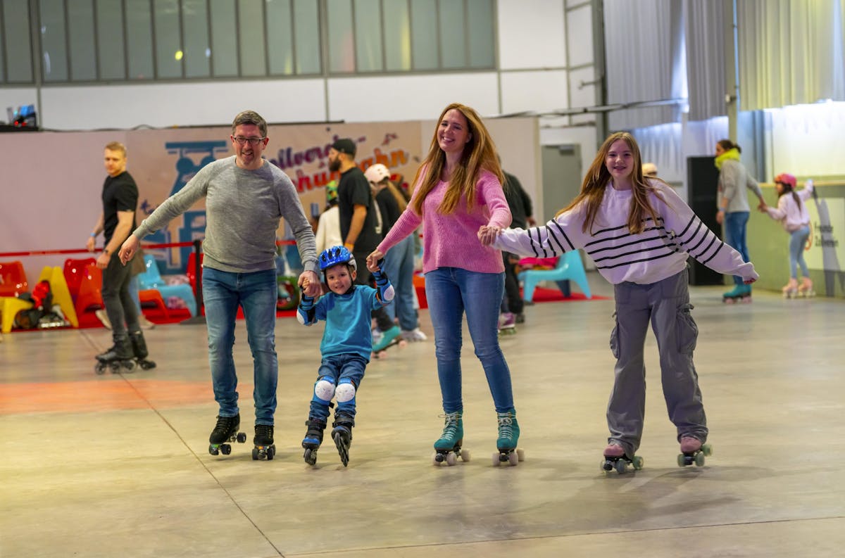 Eine fröhliche Familie fährt gemeinsam auf Rollschuhen in einer bunten Rollschuhhalle. Ein Mann, eine Frau und ein Mädchen halten einen kleinen Jungen mit Helm und Schutzausrüstung an den Händen, während dieser mit angehobenen Füßen zwischen ihnen schwebt. Im Hintergrund fahren weitere Kinder und Erwachsene auf Rollschuhen, bunte Stühle und eine bemalte Wand mit der Aufschrift Zollverein Rollschuhbahn sind sichtbar. Die Szene strahlt Freude, Bewegung und gemeinsames Freizeitvergnügen aus.