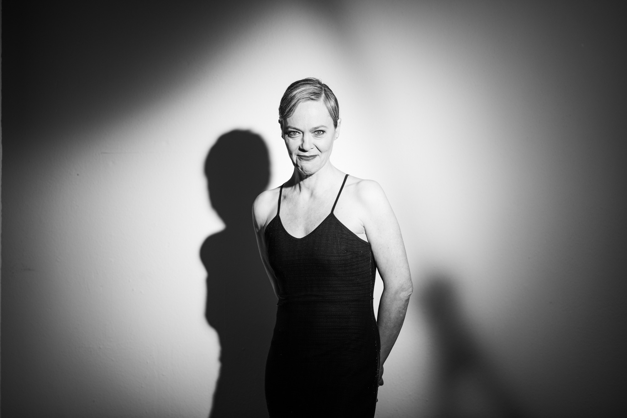 Schwarz-weiß-Porträt einer Frau mit kurz geschnittenem Haar, die in einem schlichten, ärmellosen Kleid mit Spaghettiträgern vor einer hellen Wand steht. Sie blickt lächelnd in die Kamera, die Arme hinter dem Rücken verschränkt. Durch stark gerichtetes Licht entsteht ein markanter Schatten ihres Oberkörpers an der Wand. Die Stimmung des Bildes ist elegant und stilisiert.