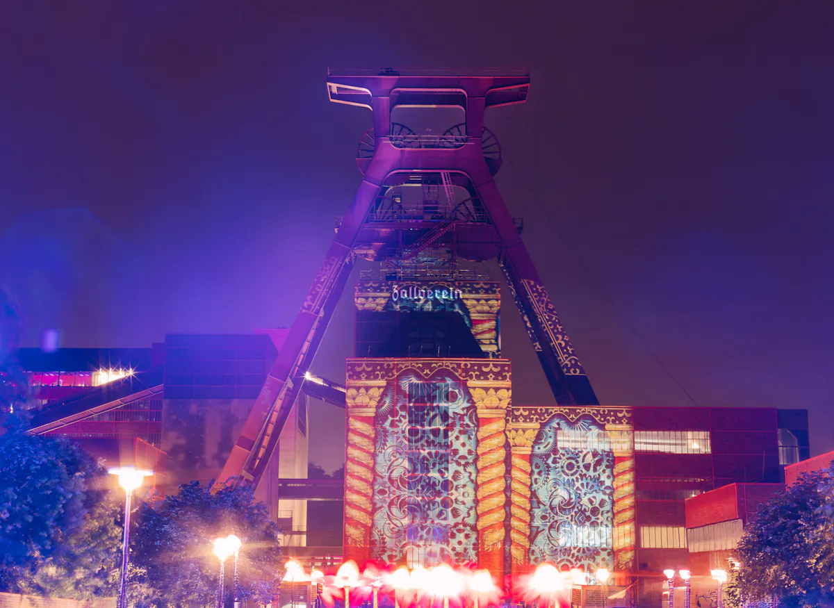 Ein ehemaliger Förderturm mit der Aufschrift Zollverein ist nachts farbenfroh angestrahlt. Auf das zentrale Gebäude darunter sind kunstvolle, bunte Lichtprojektionen mit floralen und ornamentalen Mustern geworfen. Die Szenerie ist in violettes und rotes Licht getaucht, im Vordergrund leuchten Straßenlaternen und verschwommene Lichter von Besucherinnen und Besuchern erzeugen eine lebendige Atmosphäre. Die Kulisse wirkt wie Teil einer Lichtkunstveranstaltung oder eines Festivals auf dem Gelände der Zeche Zollverein.