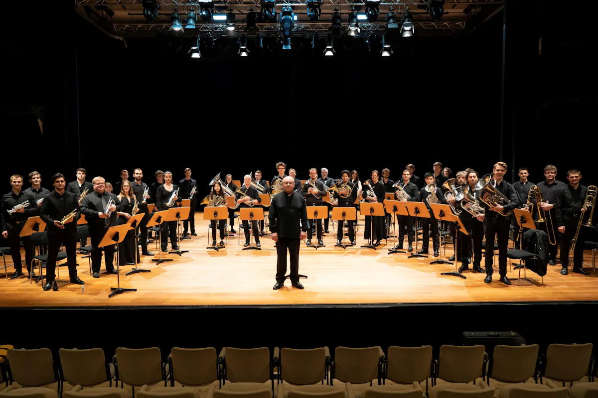 Ein großes Bläserensemble steht auf einer hell ausgeleuchteten Bühne in einem Konzertsaal. Die Musikerinnen und Musiker tragen schwarze Kleidung und halten ihre Instrumente, darunter Trompeten, Posaunen, Hörner und Tuben. In der Mitte der Bühne steht der Dirigent leicht vor dem Ensemble. Im Vordergrund sind leere Stuhlreihen des Zuschauerraums zu sehen. Die Szene wirkt feierlich und professionell, als würde das Ensemble sich vor oder nach einem Konzert präsentieren.