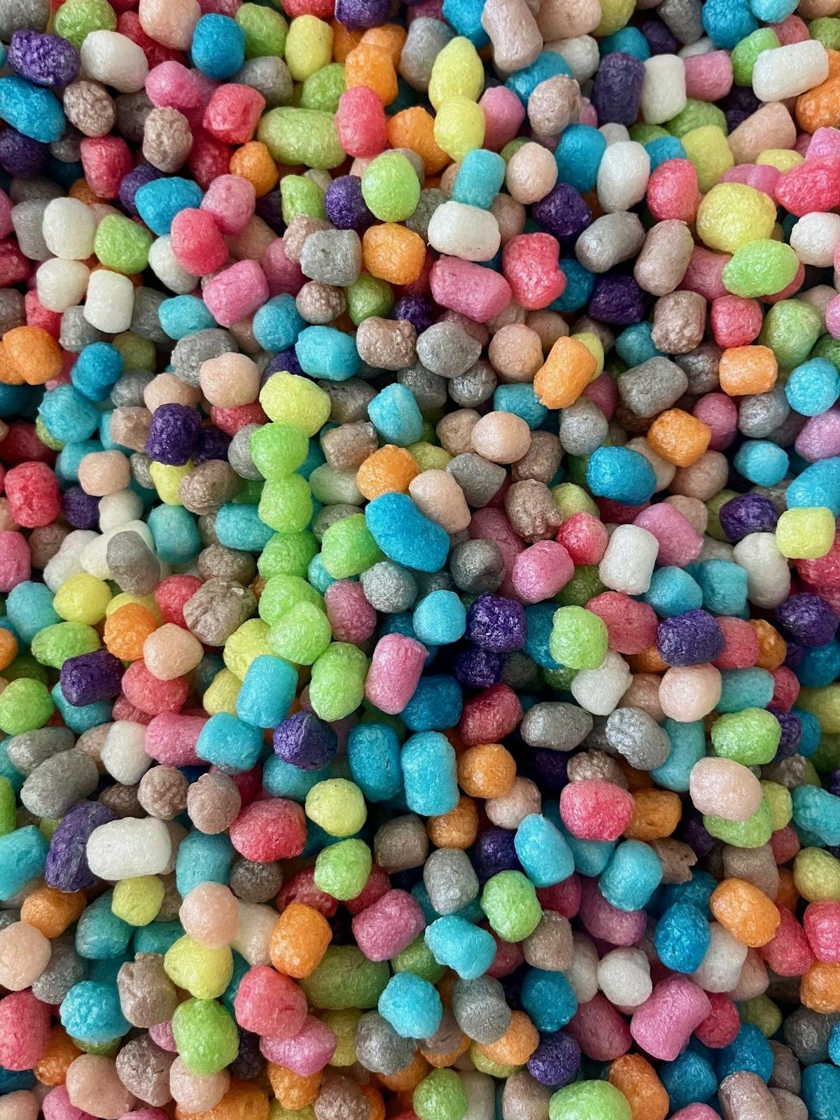 Nahaufnahme einer großen Menge kleiner, bunter, erdnussförmiger Bausteine aus Maisstärke. Sie sind in vielen verschiedenen Farben zu sehen, darunter Blau, Rosa, Gelb, Grün, Lila, Orange, Weiß und Grau. Die Oberfläche der Bausteine ist leicht glänzend und strukturiert. Sie liegen dicht an dicht und füllen das gesamte Bild aus. Die Bausteine werden häufig für kreatives Basteln verwendet und können durch Wasser aneinandergeklebt werden.