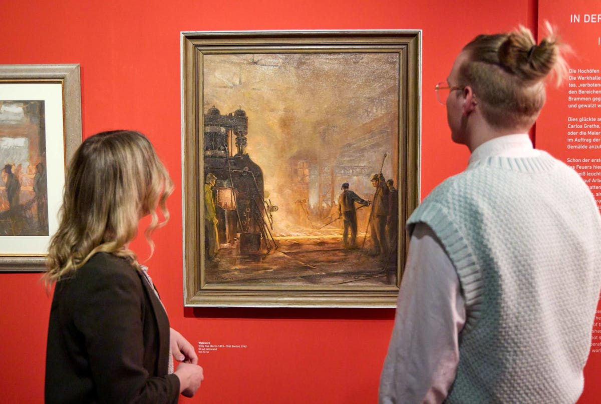 Zwei Personen stehen in einem Museum vor einem großformatigen Gemälde, das eine industrielle Szene mit Arbeitern in einer Fabrik oder Stahlhütte zeigt. Die Darstellung ist von warmen, feurigen Farben geprägt, mit hell leuchtendem Schmelzofen und rauchiger Atmosphäre. Die Ausstellung hängt vor einer intensiv roten Wand, die das Thema Industriearbeit betont. Die beiden Betrachtenden sind von hinten zu sehen und wirken konzentriert. Neben dem Bild ist ein Text zur Ausstellung angebracht, der historische Kontexte erläutert. Links ist ein weiteres, kleineres Gemälde im gleichen Stil zu sehen.