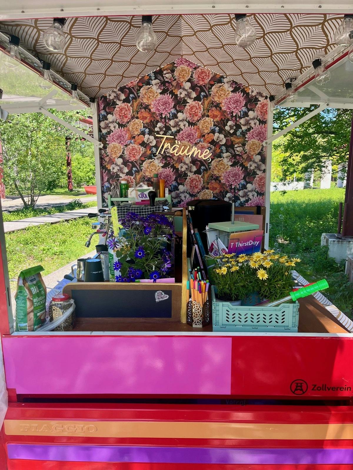 Eine bunte Präsentationsfläche mit floralem Hintergrund aus großen, farbenfrohen Blumen und dem Schriftzug Träume in leuchtender Schrift auf einem Dreirad montiert. Die Fläche ist mit vielen kleinen Gegenständen dekoriert, darunter Blumentöpfe mit violetten und gelben Blumen, buntes Schreibmaterial, kleine Bücher und Lebensmittelverpackungen. Das Dach ist mit einem gold-weißen Muster verziert und es hängen Glühbirnen an der Decke. Die Theke ist in Pink- und Rottönen gestrichen, mit dem Logo von Zollverein auf der rechten Seite. Im Hintergrund ist grüne Natur mit Bäumen und Wiese sichtbar.