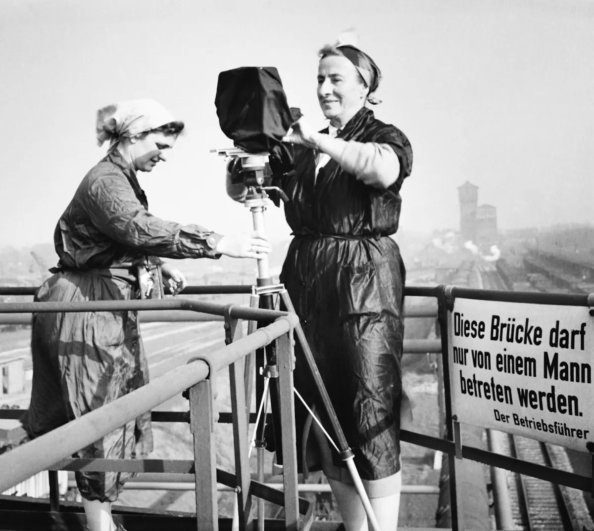 Schwarz-weiß-Fotografie von zwei Frauen in Arbeitskleidung, die auf einer Metallbrücke ein großes Kamerastativ aufbauen. Im Hintergrund ist eine Industrieanlage mit Schornsteinen zu sehen. Rechts im Bild hängt ein Schild mit der Aufschrift: Diese Brücke darf nur von einem Mann betreten werden. Der Betriebsführer. Die Szene wirkt wie ein stiller Protest gegen das Verbot und zeigt Frauen in einer typischen Männerdomäne der damaligen Zeit.
