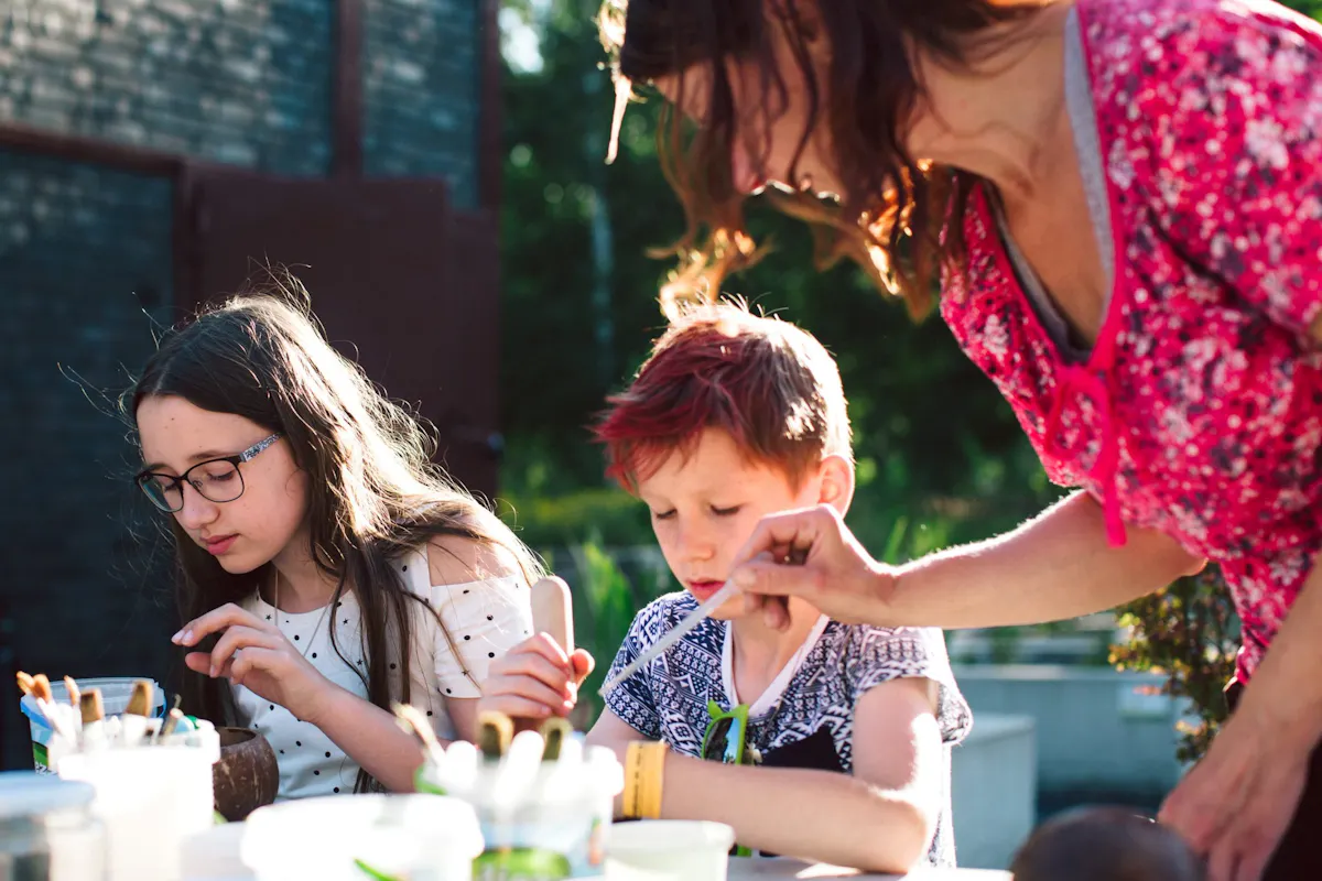 Zwei Kinder und eine Frau sitzen im Freien an einem Tisch und sind in eine kreative Tätigkeit vertieft. Das Mädchen links trägt eine Brille und ein weißes Oberteil mit schwarzen Punkten und konzentriert sich auf ihre Arbeit. Der Junge in der Mitte hat rot gefärbte Haare und bemalt konzentriert einen kleinen Gegenstand. Die Frau rechts, vermutlich eine Betreuerin, trägt ein pink gemustertes Oberteil und zeigt dem Jungen mit einem Pinsel etwas. Auf dem Tisch stehen verschiedene Pinsel, Becher und Bastelmaterialien. Die Szene spielt sich bei sonnigem Wetter im Freien ab, vermutlich bei einem Kreativworkshop oder Ferienprogramm.