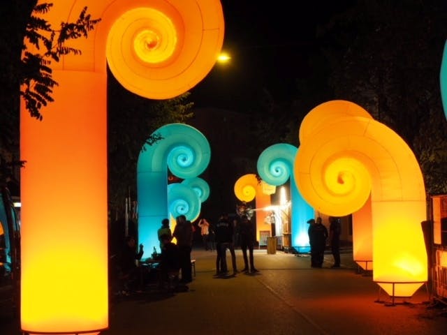 Beleuchtete, spiralförmige Skulpturen in Orange und Blau säumen einen nächtlichen Weg bei einer Outdoor-Veranstaltung. Die leuchtenden Elemente ragen hoch auf und erinnern an kunstvolle, geschwungene Säulen oder Pflanzenformen. Im Vordergrund und entlang des Weges stehen oder gehen Menschen, teils im Gespräch oder fotografierend. Die Szene ist farbenfroh, fantasievoll und vermittelt eine festliche, kunstvolle Atmosphäre im Rahmen eines Licht- oder Kulturfestivals.